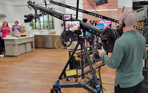 A QVC production team member using a camera to capture 3 personalities in frame. They are on a kitchen set.