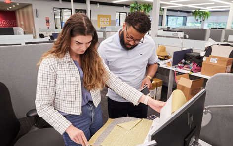 Two QVC team members with attentive looks are browsing through fabric samples at a desk. There are boxes in the background.