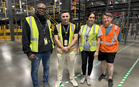 Four people wearing safety vests stand in a QVC warehouse. Three are in yellow vests and one is in an orange vest. They are smiling and there is industrial equipment in the background.