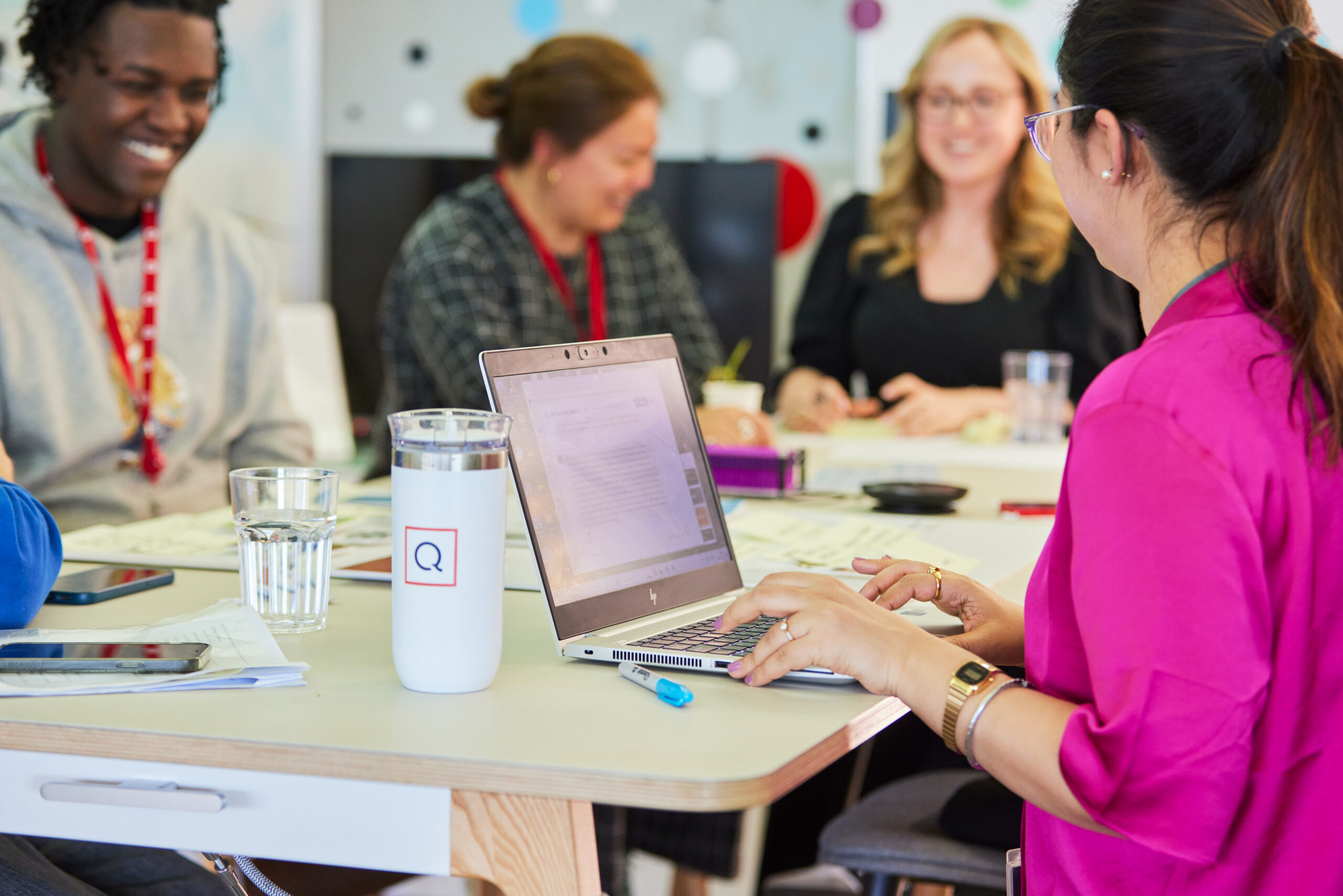 A group of four QVC team members sit around a table in a meeting room, engaged in discussion. One person in the foreground types on a laptop. A tumbler and water glass are on the table. Everyone appears to be smiling and interacting positively.