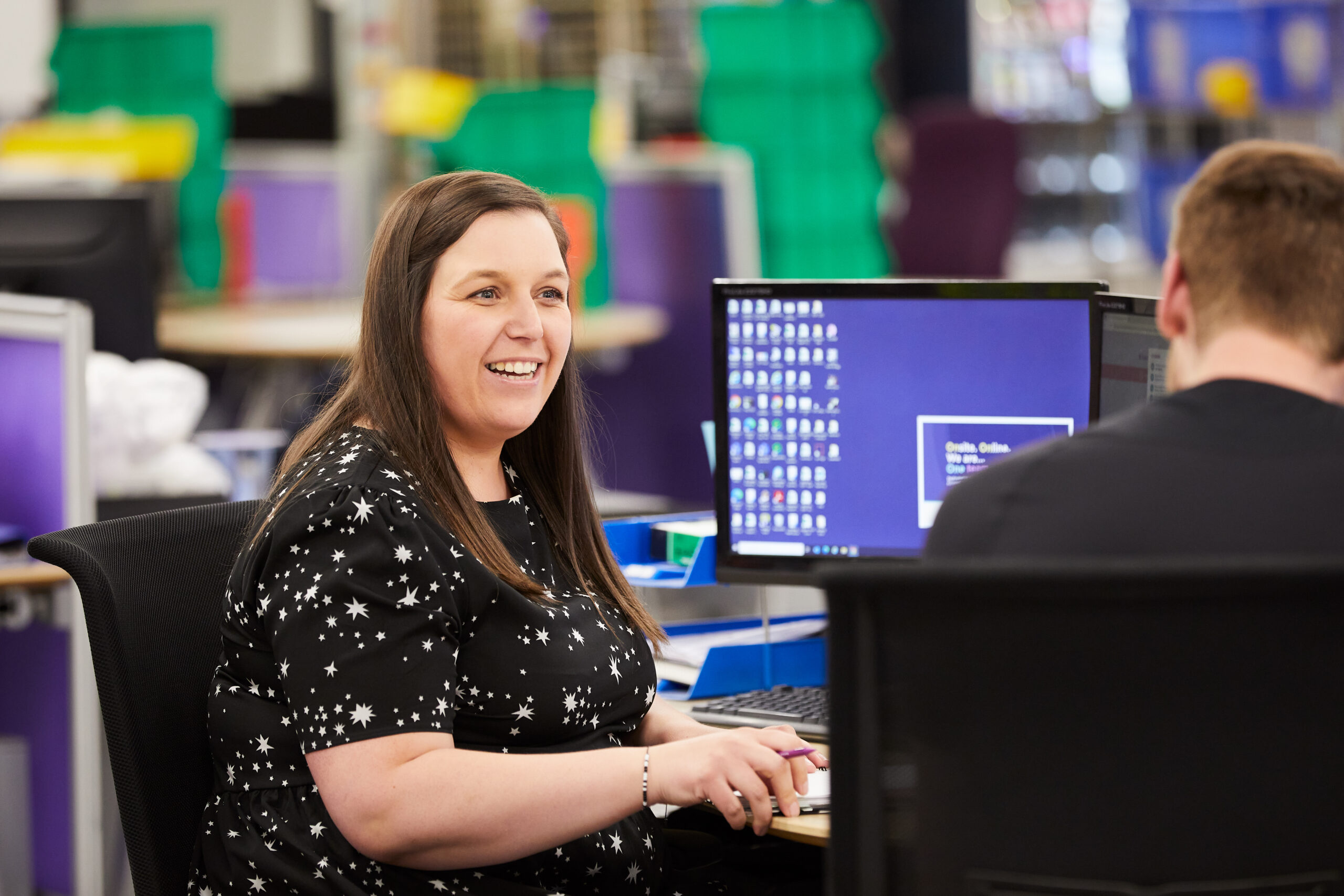 A woman is smiling at her QVC coworker, who has his back toward the camera. The woman is wearing a black shirt with stars on it and is holding a notebook and pen in front of a computer screen.