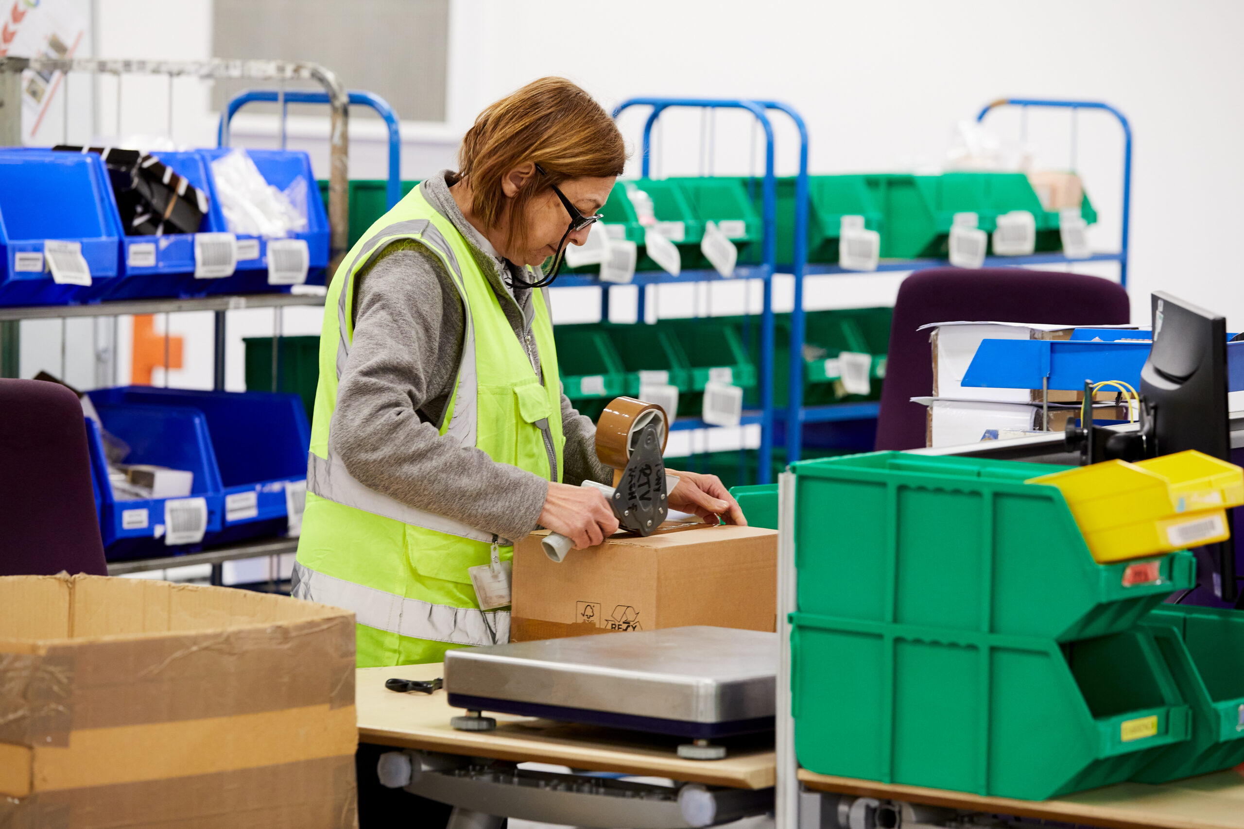 A QVC fulfillment center team members taping up a package. She is in a yellow vest and is surrounded by green and blue distribution bins.