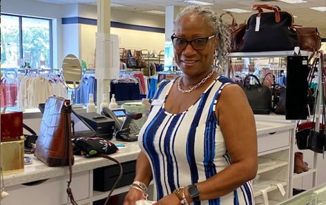 A QVC team member with gray hair and glasses stands smiling behind the QVC retail store counter, wearing a striped dress. The store interior has clothes and accessories on display in the background.