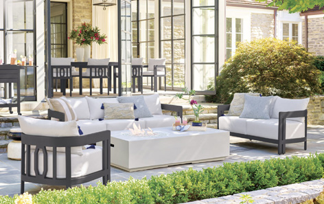 A modern outdoor patio features a white sectional sofa and two armchairs around a white fire pit table. The area is surrounded by greenery, with a stone wall and glass doors in the background, and a dining area is visible on a raised platform.