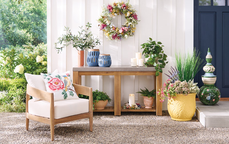 A cozy outdoor seating area features a wooden chair with a floral cushion, a table with lanterns and candles, potted plants, and a colorful wreath on a white wall. Lush greenery surrounds the space, creating a peaceful atmosphere.