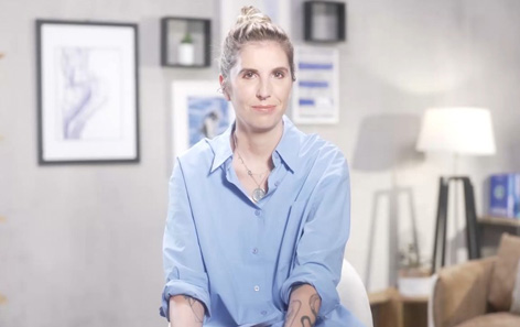 A woman with short blonde hair in a blue button-down shirt faces the camera with a slight smile. She is seated in a traditional QVC living room.