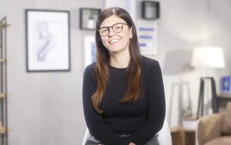 A woman with long dark hair and glasses stares is smiling into the camera. She is in a living room with framed photos and a lamp in the background. She works for QVC