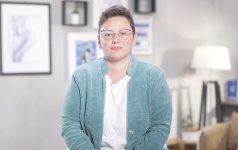 Person with short hair and glasses, wearing a teal jacket, is sitting in a cozy living room with framed pictures, shelves, and a lamp in the background.