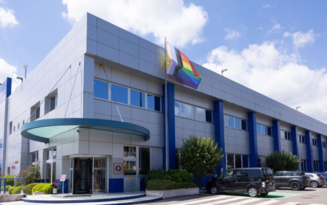 QVC's Italy modern office building with large windows, a rainbow flag, and a glass entrance. Several cars are parked nearby under a partly cloudy sky.