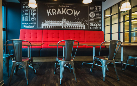 A modern cafe interior with metal chairs and red cushioned seating against a wall. The wall features a monochrome illustration and text, including Greetings from Krakow. Hanging lights illuminate the space, creating a cozy in the QVC office.