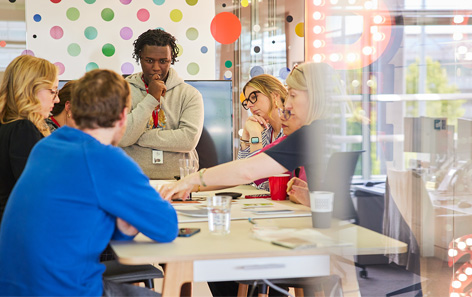 A diverse team of six QVC team members engaged in a meeting in a colorful office space. They are gathered around a table with documents and a cup of coffee. Bright polka dot wall and large windows in the background create a lively atmosphere.