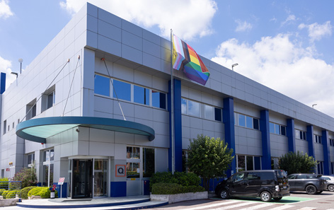 QVC's Italy modern office building with large windows, a rainbow flag, and a glass entrance. Several cars are parked nearby under a partly cloudy sky.